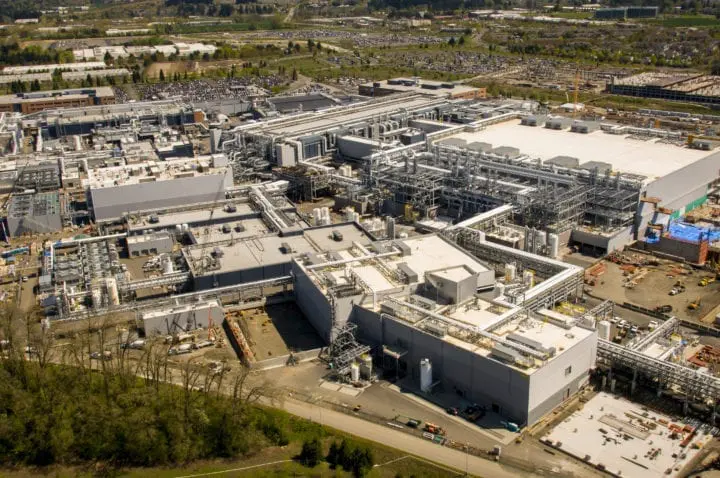Aerial view of a large industrial complex with buildings, pipes, and surrounding greenery on a sunny day.
