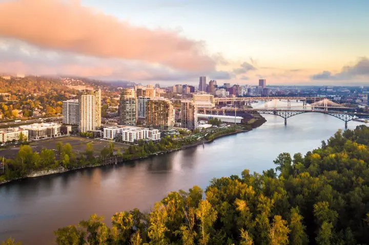 Aerial view of Portland skyline with river and bridges at sunrise, surrounded by urban and natural landscapes.