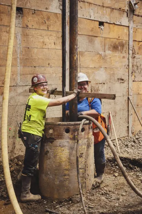 Construction workers smiling while working on a site, wearing hard hats and safety gear.