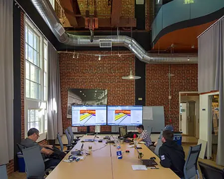 Modern conference room with brick walls, attendees at a long table viewing presentations on dual screens. Collaborative workspace setting.
