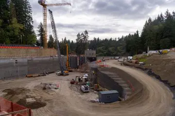 Construction site with cranes, vehicles, and equipment surrounded by trees and cloudy sky. Progress in infrastructure development.