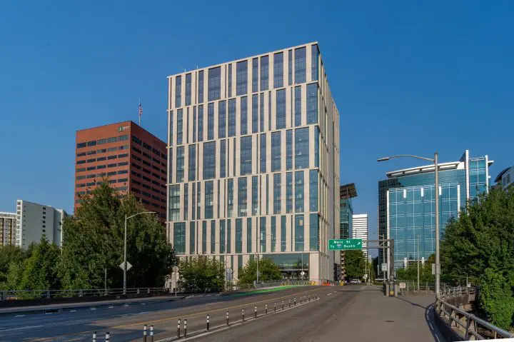 Modern cityscape with tall, glass office buildings under a clear blue sky, viewed from an empty road lined with trees.