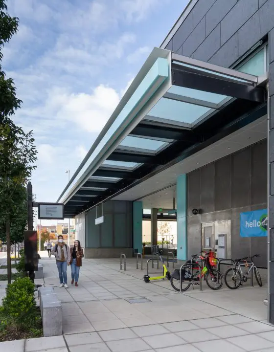 Modern transit station entrance with bike racks and e-scooters. Two people walk along the sidewalk under a sleek glass canopy.