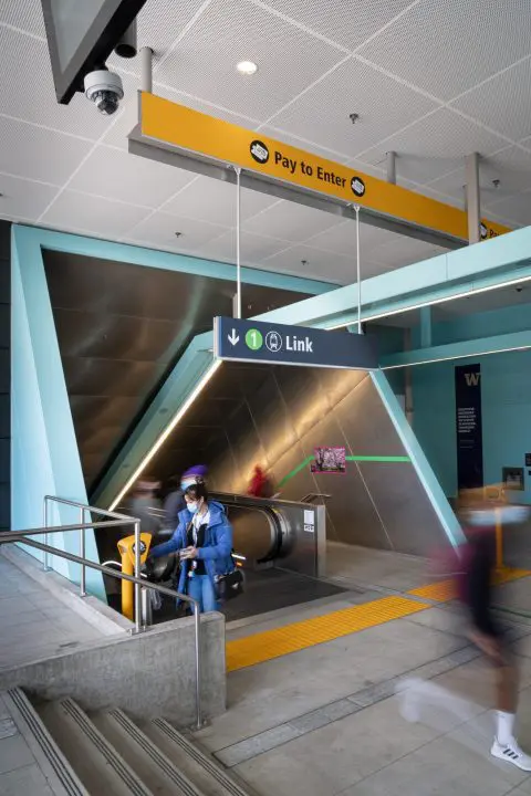 TransLink station entrance with escalator, people in motion, and Pay to Enter sign, creating a dynamic urban transit scene.