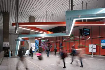 Modern transit station interior with people in motion, vibrant red accents, escalator, and signage.