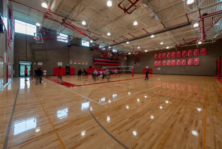 Bright gymnasium with a polished wooden floor, red decor, and people playing sports.