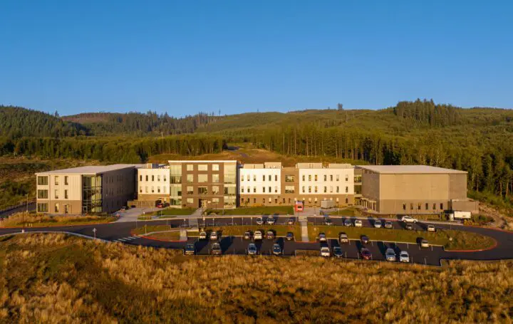 Modern building surrounded by trees and cars in parking lot, set in scenic hillside under a clear blue sky.