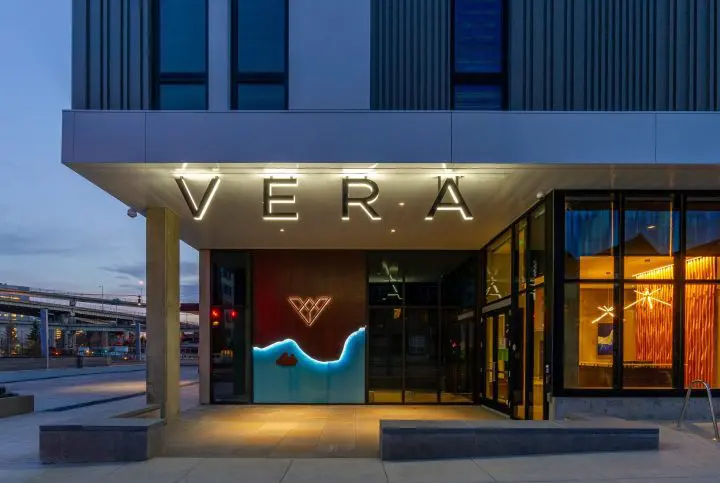 Hotel entrance with glowing Vera sign and modern decor at dusk. Glass facade displays warm, inviting interior lighting.