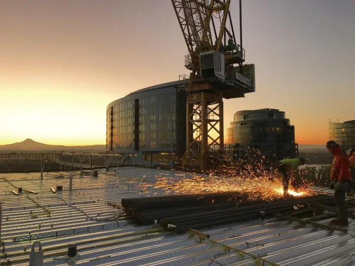 Construction workers welding on skyscraper at sunrise, sparks flying, modern buildings in background, cityscape view.