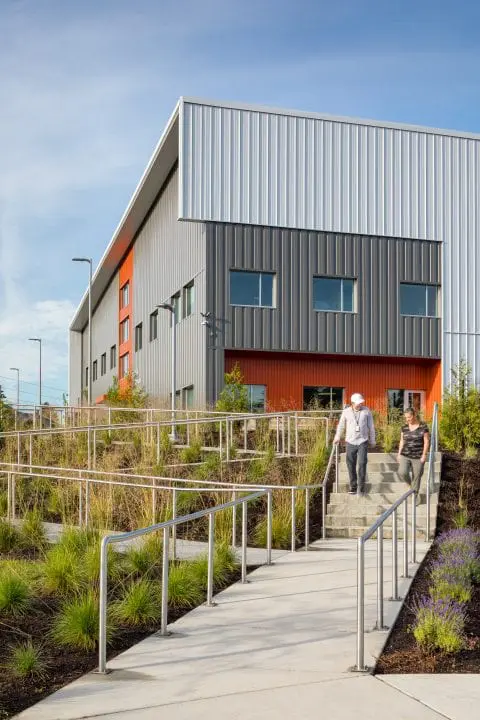 Modern industrial building with orange accents; two people walk down stairs beside landscaped pathways under a clear sky.