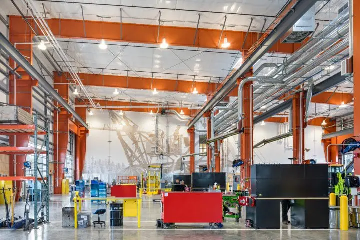 Modern industrial workshop with bright lighting, red beams, and organized workstations. High ceiling and metal pipes are visible.