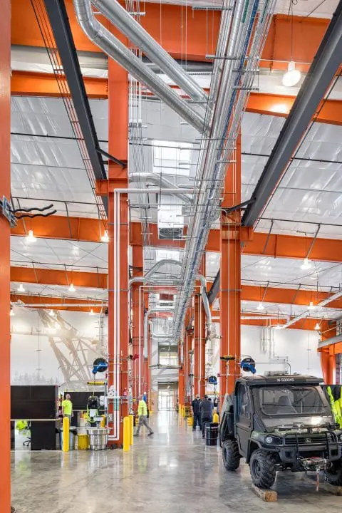 Industrial facility with orange beams and visible pipes, workers in safety gear busy with tasks, and an off-road vehicle inside.