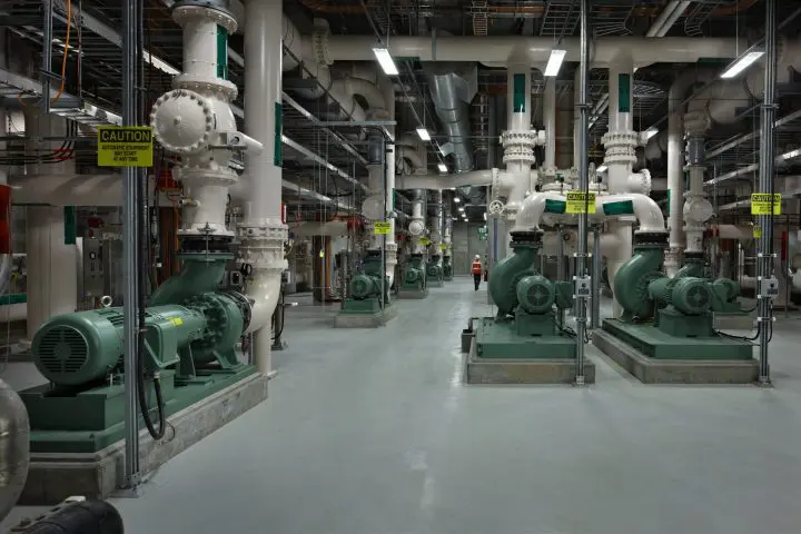 Industrial facility interior with large machinery and overhead pipes. Worker in the distance. Safety signs visible.