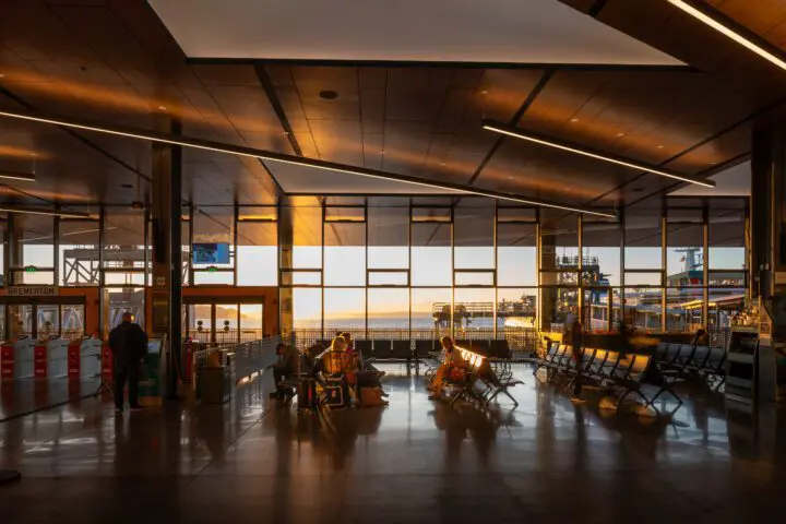 Modern ferry terminal interior at sunset with travelers, large windows, reflections on shiny floor, and view of boats outside.