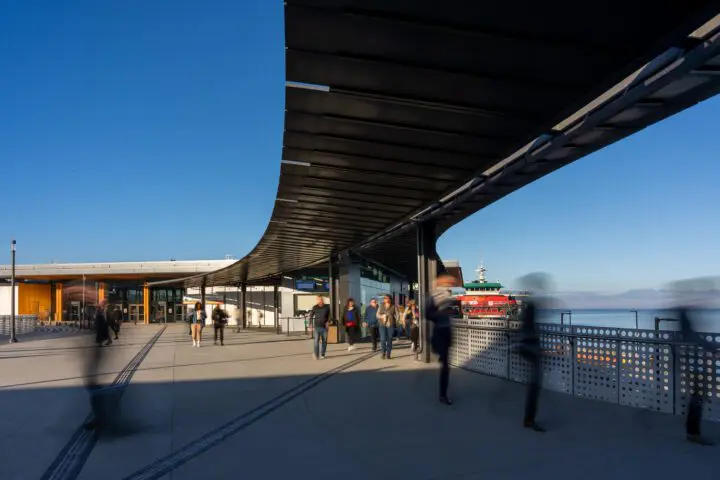 Modern waterfront terminal with people walking, ferry docked under clear blue sky.