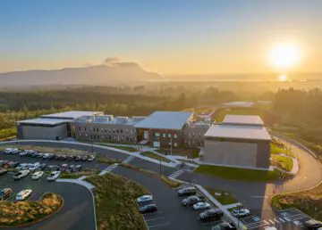 Aerial view of modern school campus at sunrise, surrounded by lush greenery, with mountains in the background and a parking lot in front.