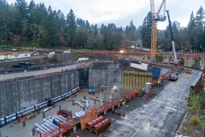 Construction site with cranes and equipment surrounded by trees. Workers busy with infrastructure project in a forested area.