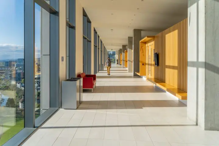 Modern hallway with large windows, natural light, and a person walking. Features red seating, yellow wooden accents, and city views.