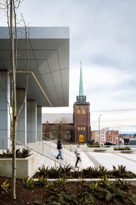 Modern building entrance with people walking, historic church in background, overcast sky. Urban contrast scene.