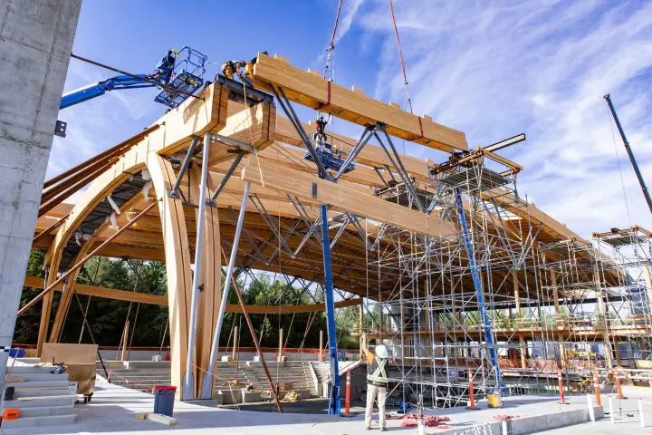 Construction workers on scaffolding and cranes build a large wooden structure under a clear blue sky.