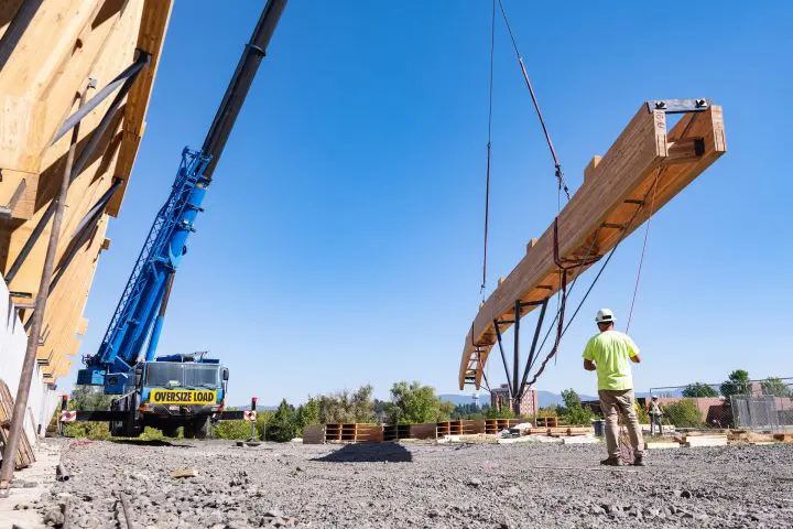 Crane lifting large wooden beam at construction site under clear blue sky, with worker guiding the operation.