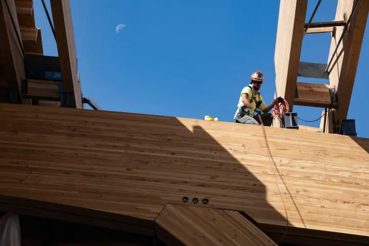Construction worker on a wooden structure under a blue sky with the moon visible, holding equipment.