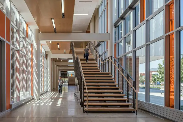Modern office interior with large windows, sunlight, and a wooden staircase. Two people walking, one up the stairs.