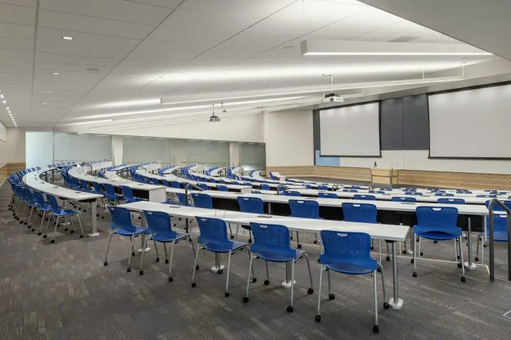 Modern lecture hall with blue chairs and white desks in a tiered arrangement, featuring dual projectors and screens.