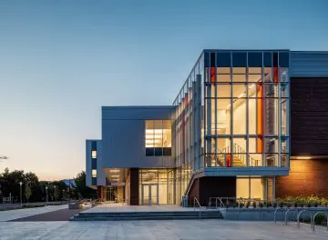 Modern building exterior with large glass windows and illuminated interior at sunset, showcasing contemporary architectural design.