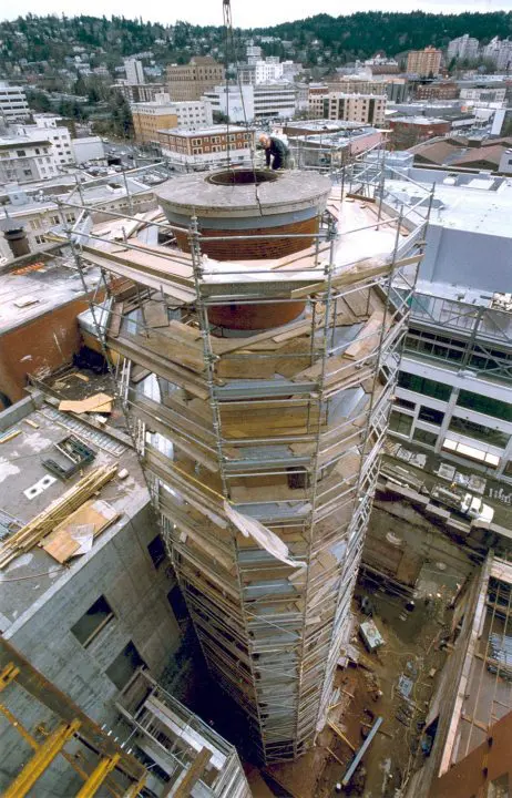 Construction worker on scaffolding around a chimney in urban setting, surrounded by cityscape and hills in the background.