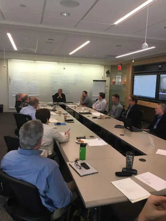 Business meeting in a conference room with diverse group discussing ideas, whiteboard notes, and laptops.