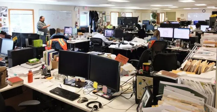Open-plan office with people working at desks, computers, and whiteboard with presentation. Busy workplace environment.