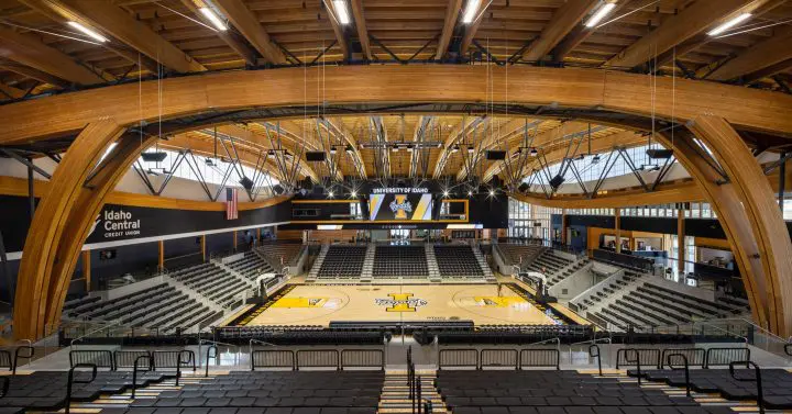 Modern basketball arena with wooden beams, empty seats, and an illuminated scoreboard; University of Idaho branding visible.
