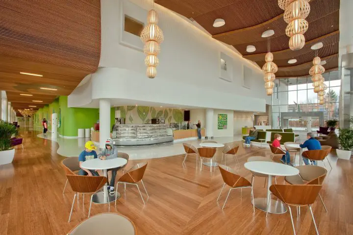 Spacious modern lobby with wooden flooring, stylish lighting, and people sitting at round tables in a bright, open setting.