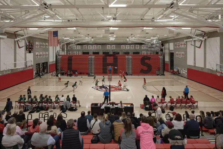 Wide shot of a high school volleyball game with players in action, a crowd in the stands, and LHS visible on the bleachers.