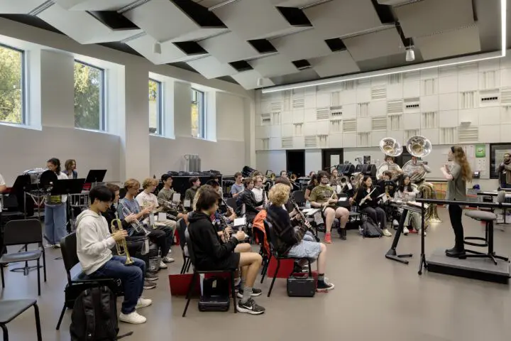 Band rehearsal in a spacious, modern room with musicians and a conductor, featuring various instruments like tubas and trumpets.