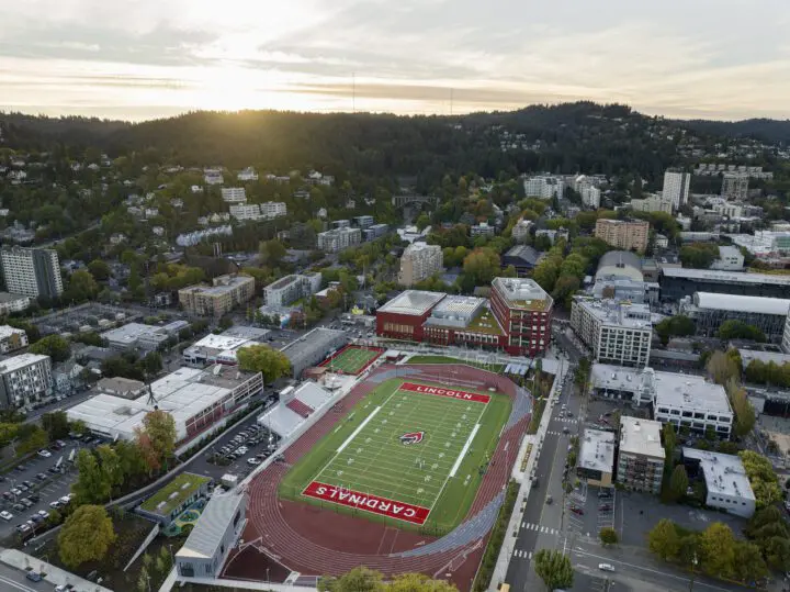 Aerial view of a school stadium amid urban landscape, surrounded by trees and buildings at sunset.