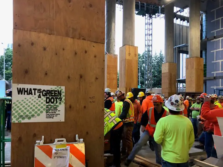 Construction workers in safety gear gather indoors near pillars, with a sign promoting anti-harassment measures on the wooden panel.