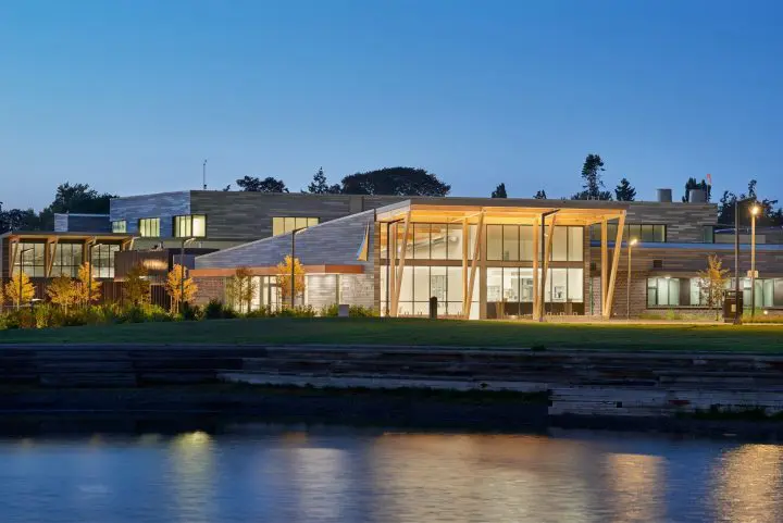Modern building with large windows illuminated at dusk, surrounded by greenery and reflecting on a calm waterbody.