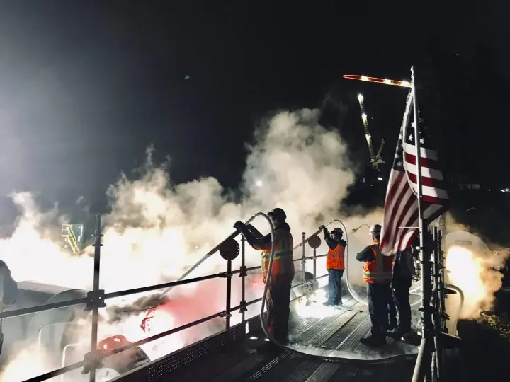 Nighttime construction site with workers in safety gear operating hoses, surrounded by steam, under an American flag.