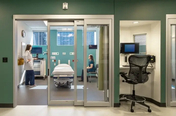 Modern medical exam room with healthcare professional and patient, featuring a hospital bed, computer station, and green walls.