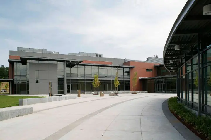 Modern building exterior with large windows and spacious courtyard, surrounded by greenery under a cloudy sky.