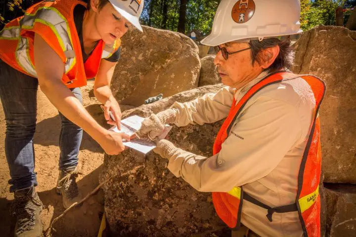 Construction workers discussing plans at a rocky site, wearing safety gear and helmets, collaborating on project details outdoors.