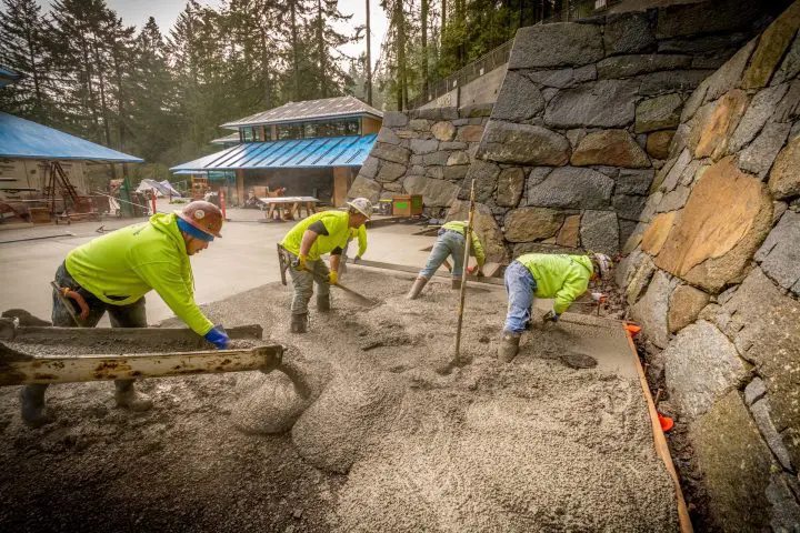 Construction workers pouring and smoothing concrete near a stone wall, outdoor site with forested background.