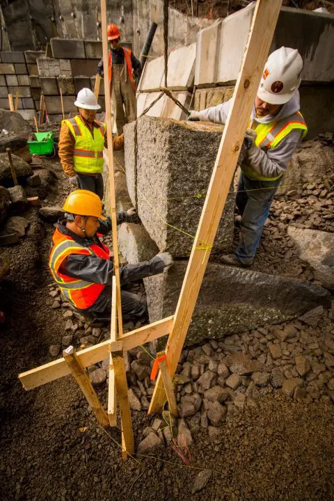 Construction crew wearing safety gear installs large stone with wooden support at building site.