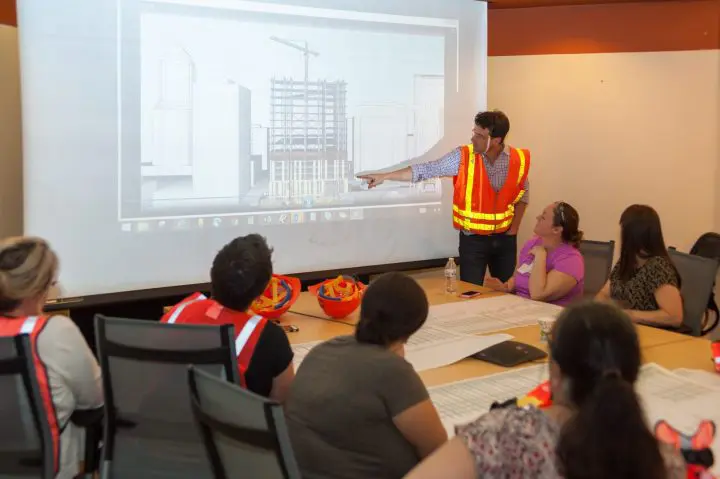 Construction team reviewing building plans on projector, wearing safety vests in a meeting room.