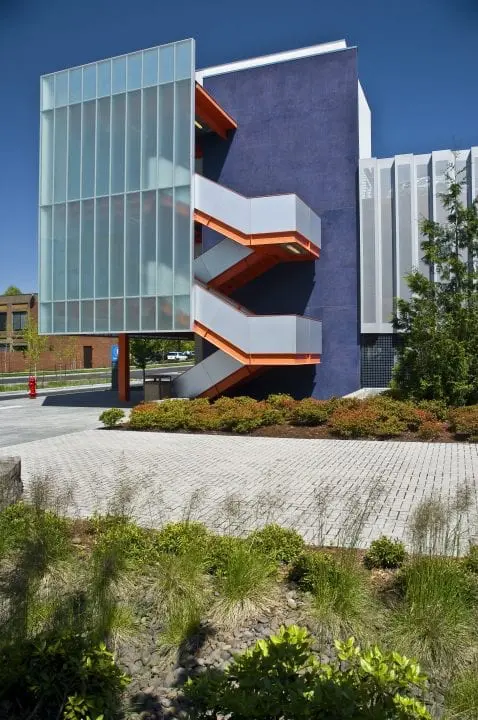 Modern building with bold blue and orange exterior, featuring a striking zig-zag staircase and lush landscaping in foreground.