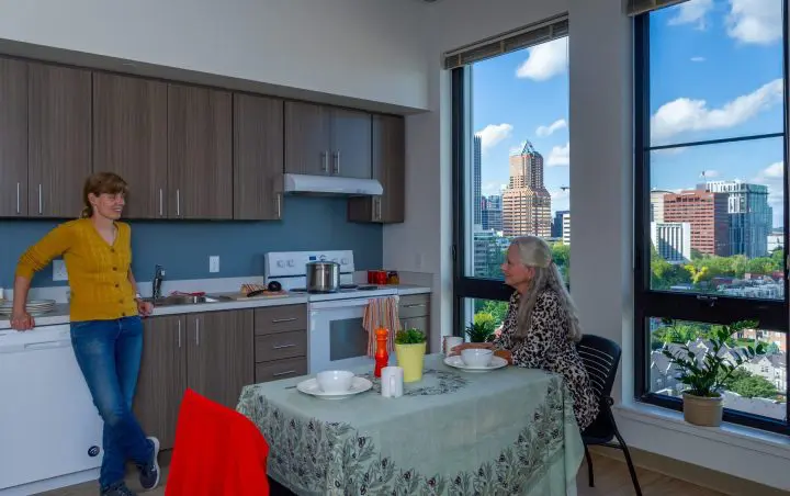 Two people chatting in a modern, sunlit kitchen with cityscape views through large windows.