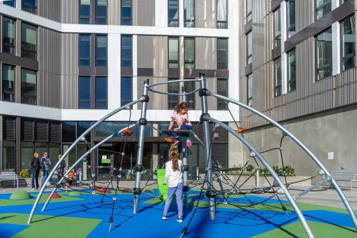 Children playing on a climbing structure in a modern playground with a building in the background. Sunny day and vibrant colors.