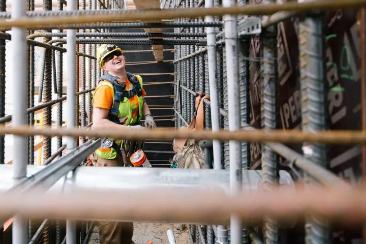 Construction worker smiling while building structure, wearing a hard hat and safety vest on site.